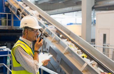 a stock image of a worker who's wearing a high vis jacket, a white safety helmet and safety goggles, who's speaking on a walkie talkie and is standing in a recycling plant.